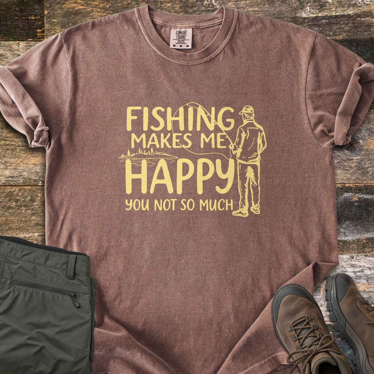 Fishing Makes Me Happy Comfort Colors T-shirt in Espresso, laid flat. Pale yellow print of a man in a cap casting a fishing rod by a tree-lined lake, with the words Fishing Makes Me Happy You Not So Much. Laid on weathered wood with olive pants and brown hiking boots.