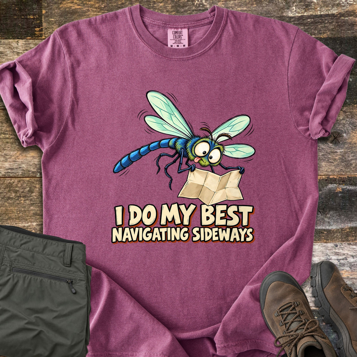 I Do My Best Navigating Sideways Comfort Colors T-shirt in Berry, laid flat. A wide-eyed cartoon dragonfly with its tongue out holds a folded paper map above the words I Do My Best Navigating Sideways. Olive pants and brown hiking boots lie beside it on weathered wood planks.