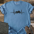 Snowmobile Heartbeat Comfort Colors T-shirt in Blue Jean, laid flat. A black heartbeat line runs across the chest with a snowmobile silhouette in its center, with no words. Sleeves are rolled, on weathered wood planks beside folded olive pants and brown hiking boots.