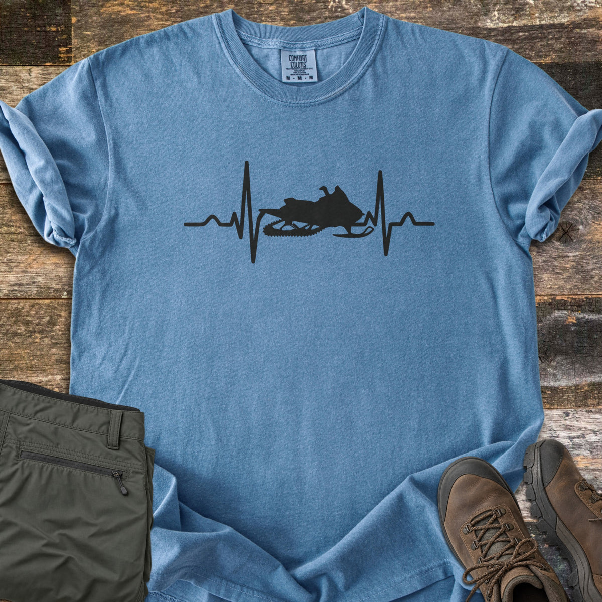 Snowmobile Heartbeat Comfort Colors T-shirt in Blue Jean, laid flat. A black heartbeat line runs across the chest with a snowmobile silhouette in its center, with no words. Sleeves are rolled, on weathered wood planks beside folded olive pants and brown hiking boots.