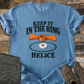 Keep It in the Ring Comfort Colors T-shirt in Blue Jean, laid flat. An orange winged helice target floats above a white ring inside a green circle, between the words Keep It in the Ring and Helice. Olive pants and brown hiking boots sit beside it on weathered wood.