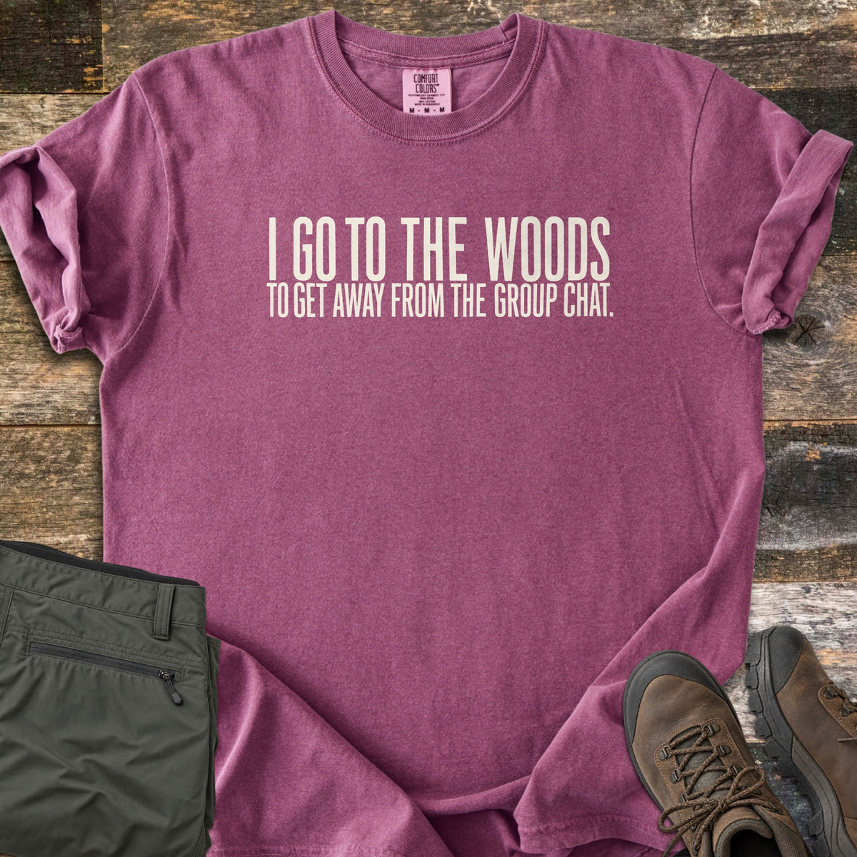 Away From The Group Chat Comfort Colors T-shirt in Berry, laid flat. Off-white block letters read I GO TO THE WOODS above TO GET AWAY FROM THE GROUP CHAT. The shirt has rolled sleeves and lies on weathered wood boards beside folded olive green pants and brown hiking boots.