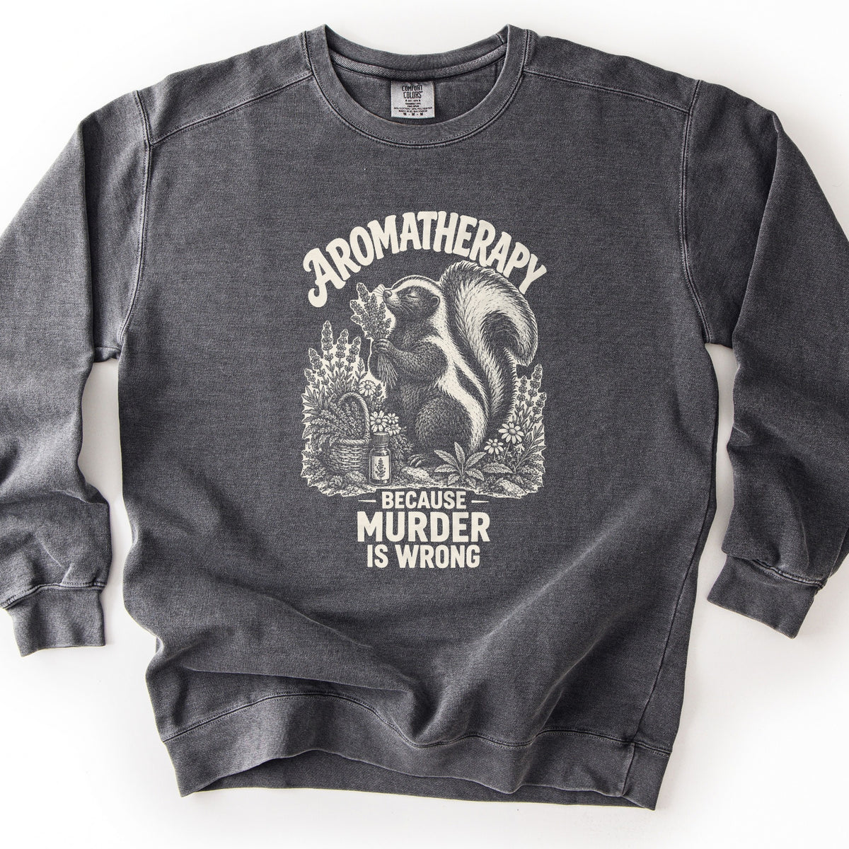 Aromatherapy Because Murder Is Wrong Comfort Colors crewneck sweatshirt in Pepper, laid flat. Cream line art of a skunk sniffing a bundle of lavender among wildflowers, a basket and an essential oil bottle, with Aromatherapy above and Because Murder Is Wrong below. Shown on a white surface.