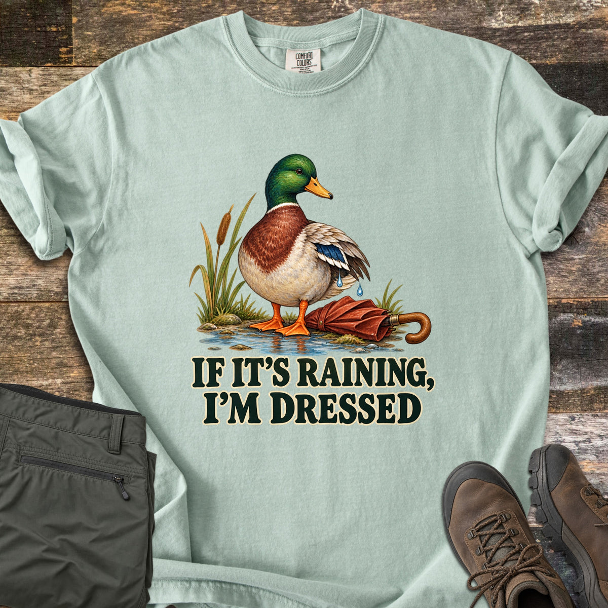 If It's Raining, I'm Dressed Comfort Colors T-shirt in Bay, laid flat. A mallard duck stands in a puddle by cattails, water dripping off its wing, next to a closed red umbrella, above the words If It's Raining, I'm Dressed in dark green. Olive pants and brown hiking boots lie on weathered wood.