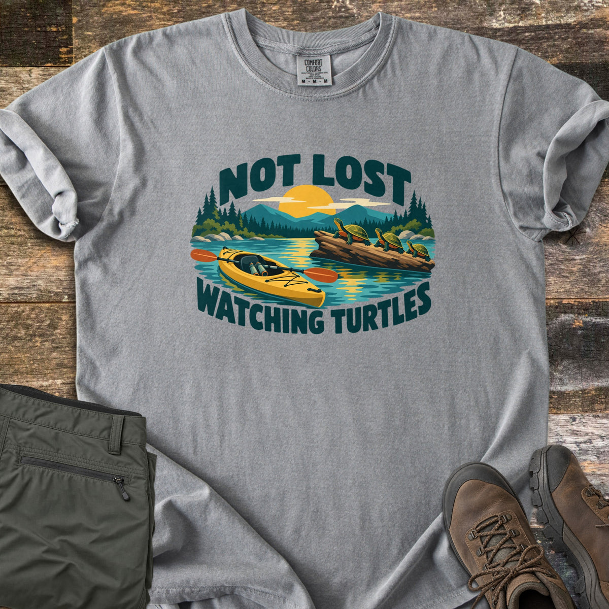 Not Lost Watching Turtles Comfort Colors T-shirt in Grey, laid flat. The words Not Lost Watching Turtles frame a lake scene of a yellow kayak and turtles sunning on a log under a setting sun and pine trees. Green pants and brown hiking boots lie beside it on weathered wood.