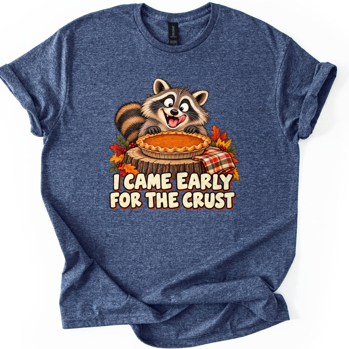 I Came Early for the Crust Comfort Colors T-shirt in Heather Navy, laid flat. A grinning cartoon raccoon licks its lips over a pumpkin pie on a tree stump with autumn leaves and a plaid napkin, above the words I CAME EARLY FOR THE CRUST. Sleeves are rolled on a white background.