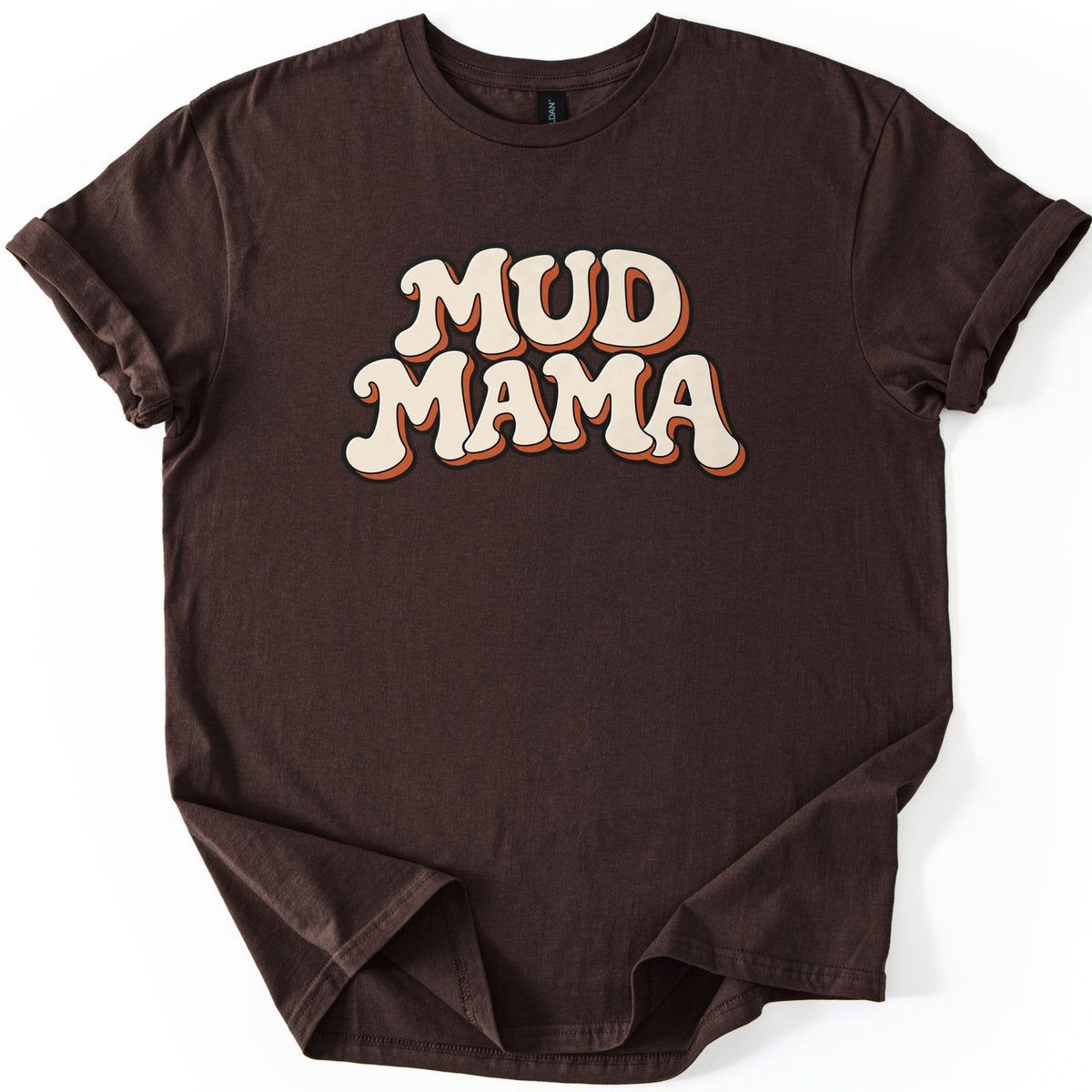 Mud Mama Comfort Colors T-shirt in Dark Chocolate, laid flat. The words MUD MAMA are printed across the chest in cream retro bubble letters with a black outline and an orange drop shadow. The shirt has rolled sleeves and lies on a white background.
