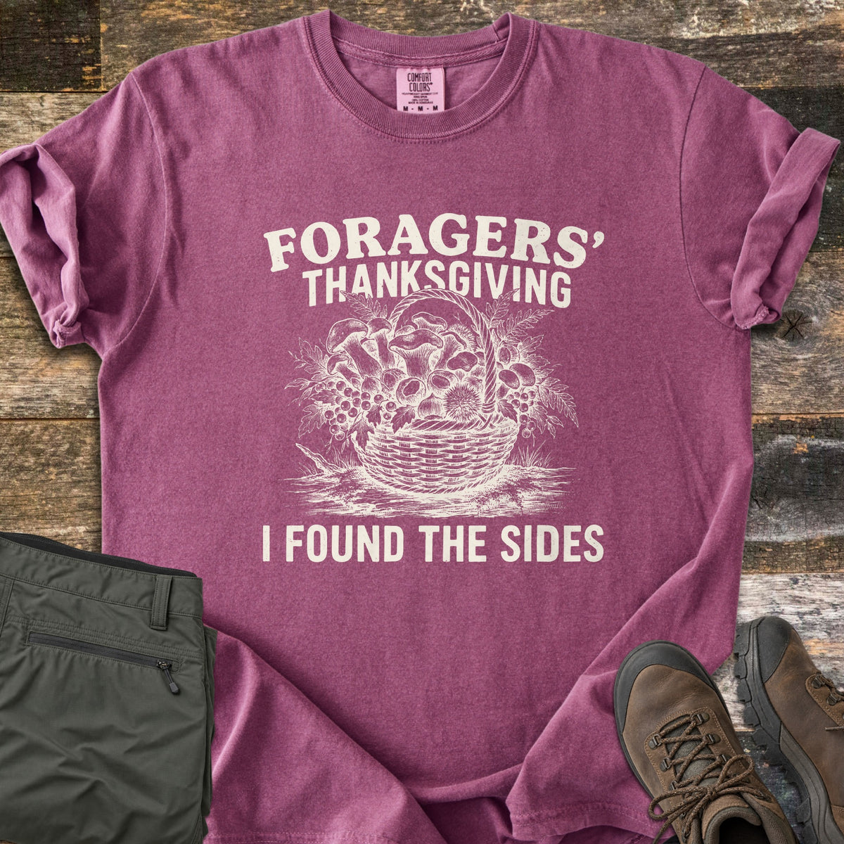 Foragers Thanksgiving Comfort Colors T-shirt in Berry, laid flat. A cream sketch of a woven basket full of mushrooms, berries and leaves sits between the words Foragers' Thanksgiving and I Found the Sides. Folded olive hiking pants and brown boots lie on weathered wood.