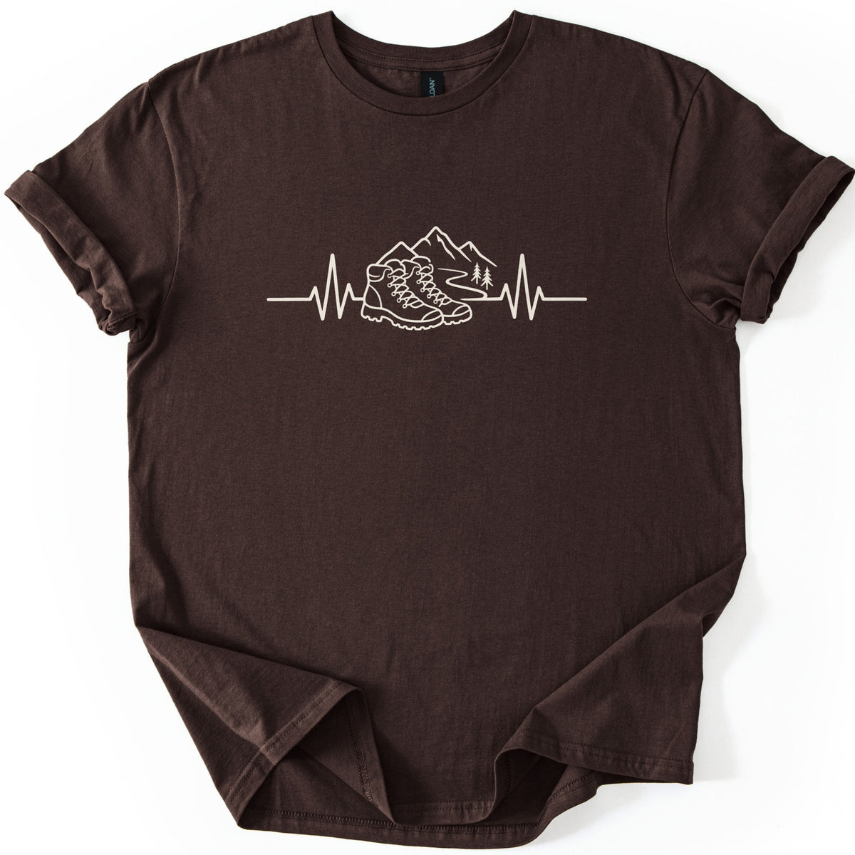 Hiking Heartbeat Comfort Colors T-shirt in Dark Chocolate, laid flat. A cream line drawing of a heartbeat line that turns into a pair of hiking boots in front of mountains, a winding trail and two pine trees; no words. Sleeves are cuffed on a white background.