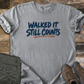 Walked It Still Counts Comfort Colors T-shirt in Grey, laid flat. Navy brush-stroke letters read Walked It Still Counts, with a dashed orange line and the small letters MTB below. Rolled sleeves, on weathered wood with olive hiking pants and brown hiking boots.