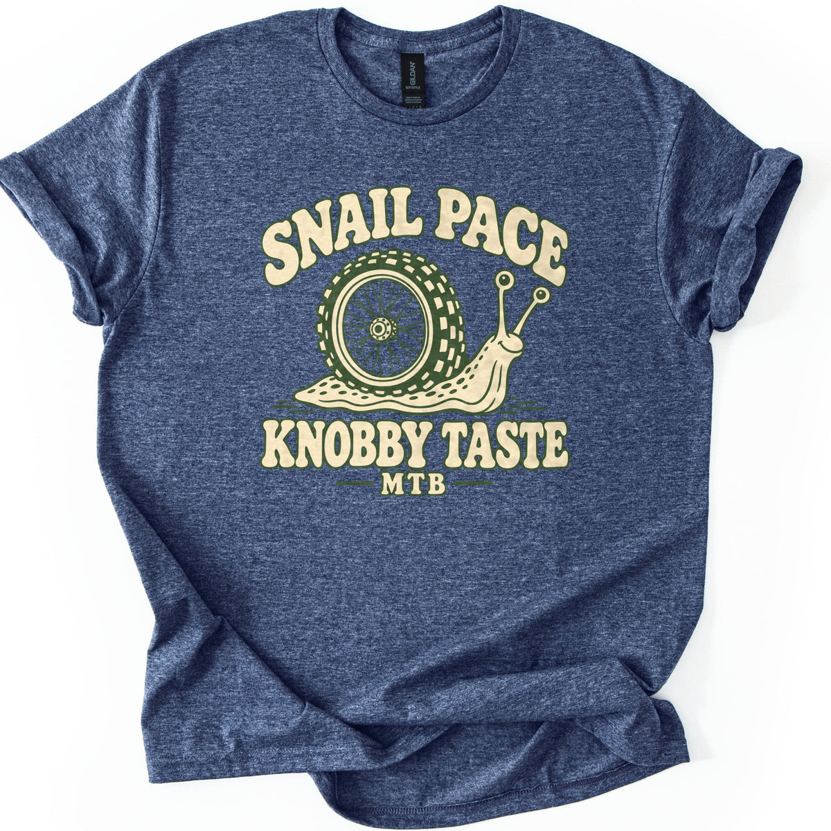 Snail Pace Knobby Taste Comfort Colors T-shirt in Heather Navy, laid flat. A smiling cartoon snail with a knobby mountain bike tire for a shell sits between the words Snail Pace and Knobby Taste, with MTB below, in cream and green. Sleeves are rolled on a white background.