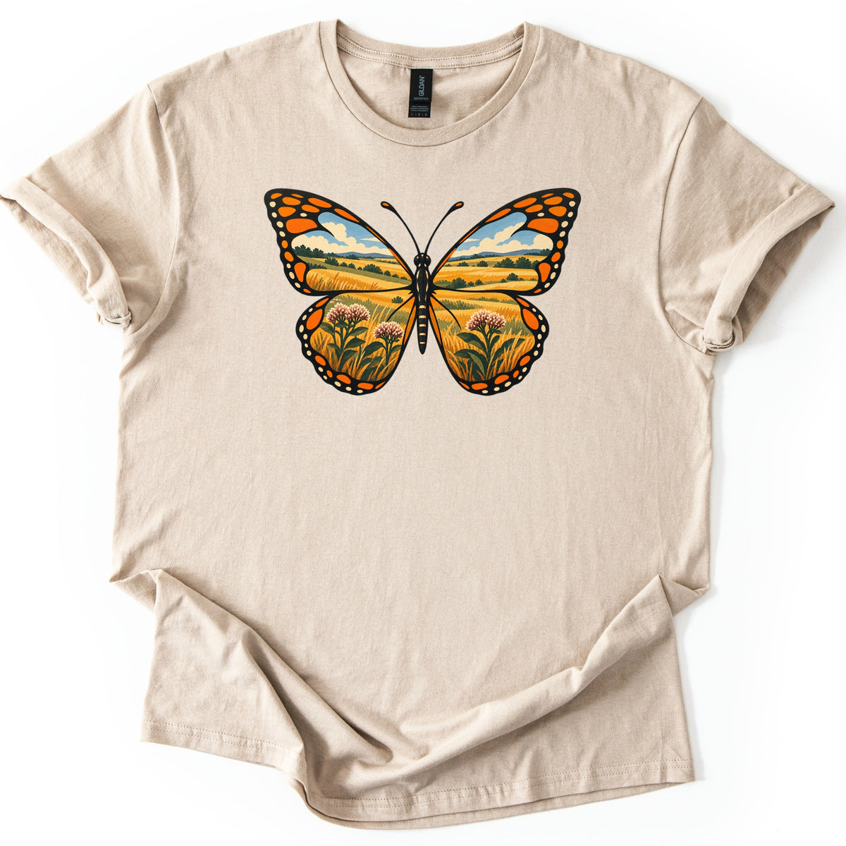 Monarch Prairie Comfort Colors T-shirt in Sand, laid flat. A monarch butterfly with orange and black wings that frame a prairie scene of golden grass, rolling hills, blue sky, and pink milkweed blooms. No words. The shirt lies on white with cuffed sleeves.