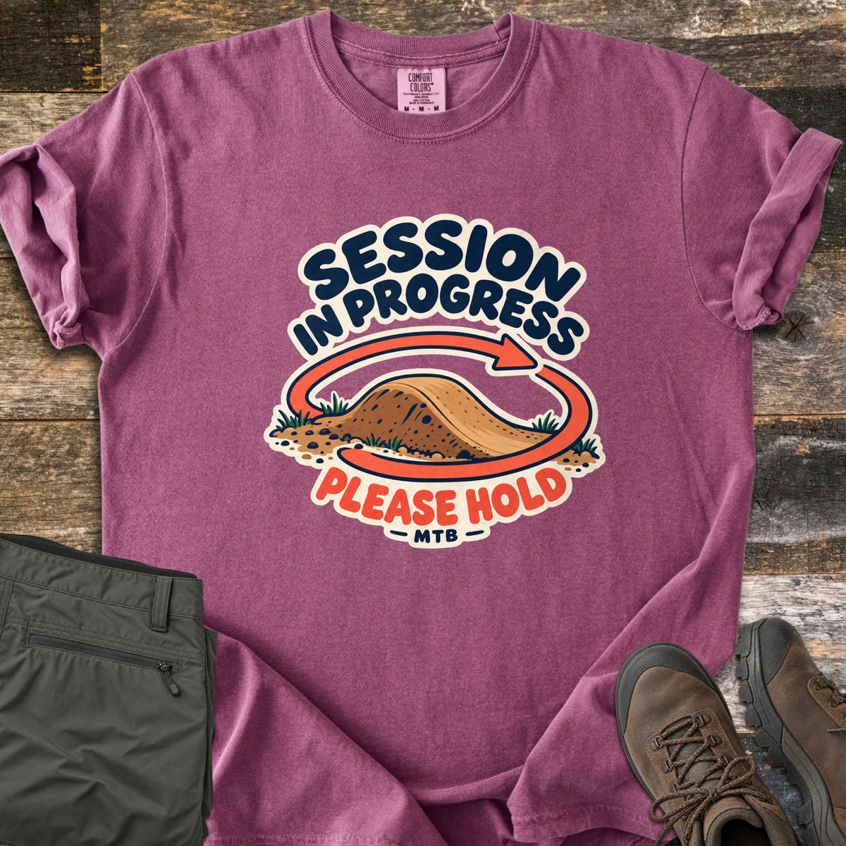 Session in Progress Comfort Colors T-shirt in Berry, laid flat. A coral arrow loops around a dirt jump between the words Session in Progress and Please Hold, with MTB below. Sleeves rolled, on weathered wood beside folded olive pants and brown hiking boots.