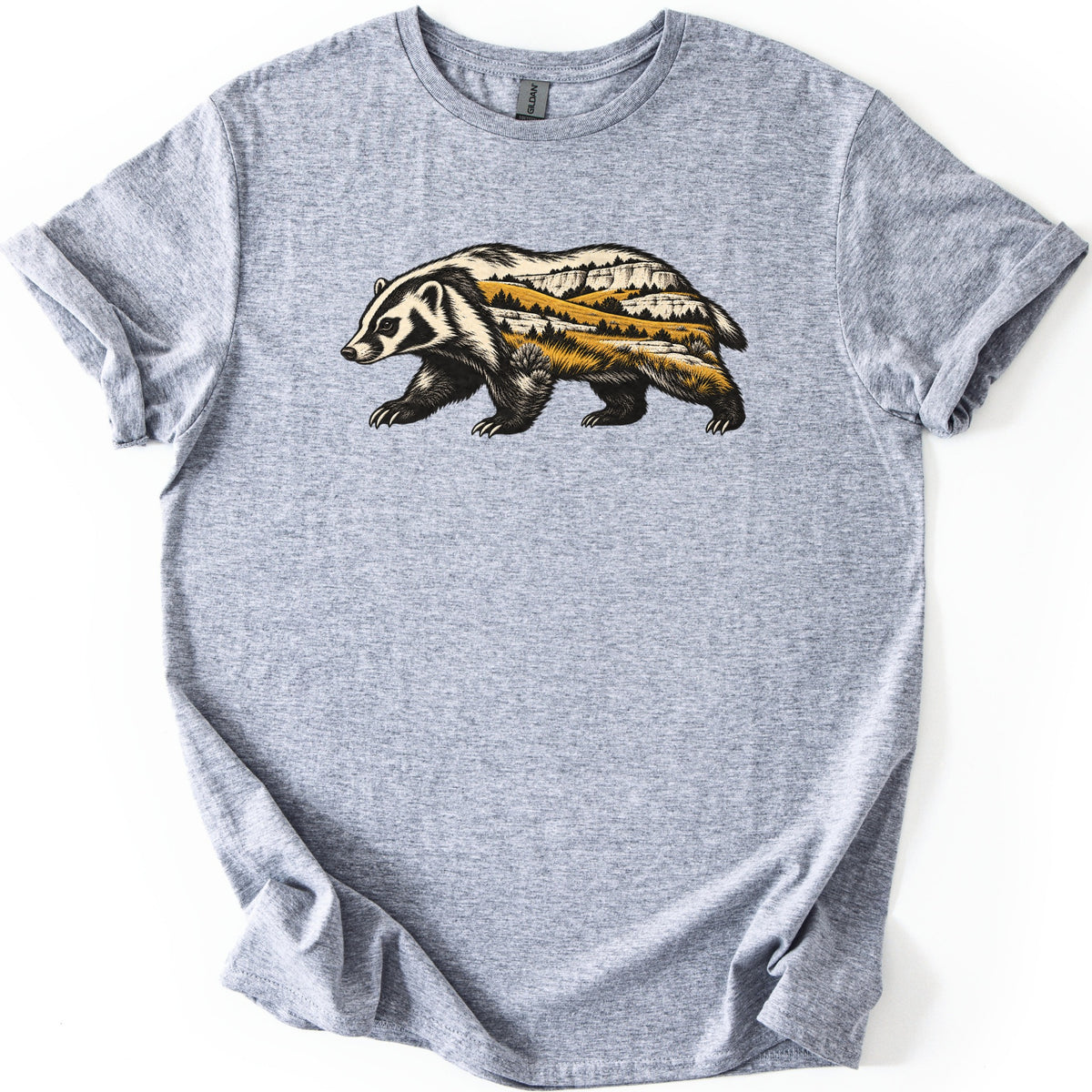 Badger Prairie Bluffs Comfort Colors T-shirt in Sport Grey, laid flat. A badger walking left, its body filled with a scene of white bluffs, golden prairie grass and dark pine trees, with no words. The shirt lies on a white surface with its sleeves cuffed.
