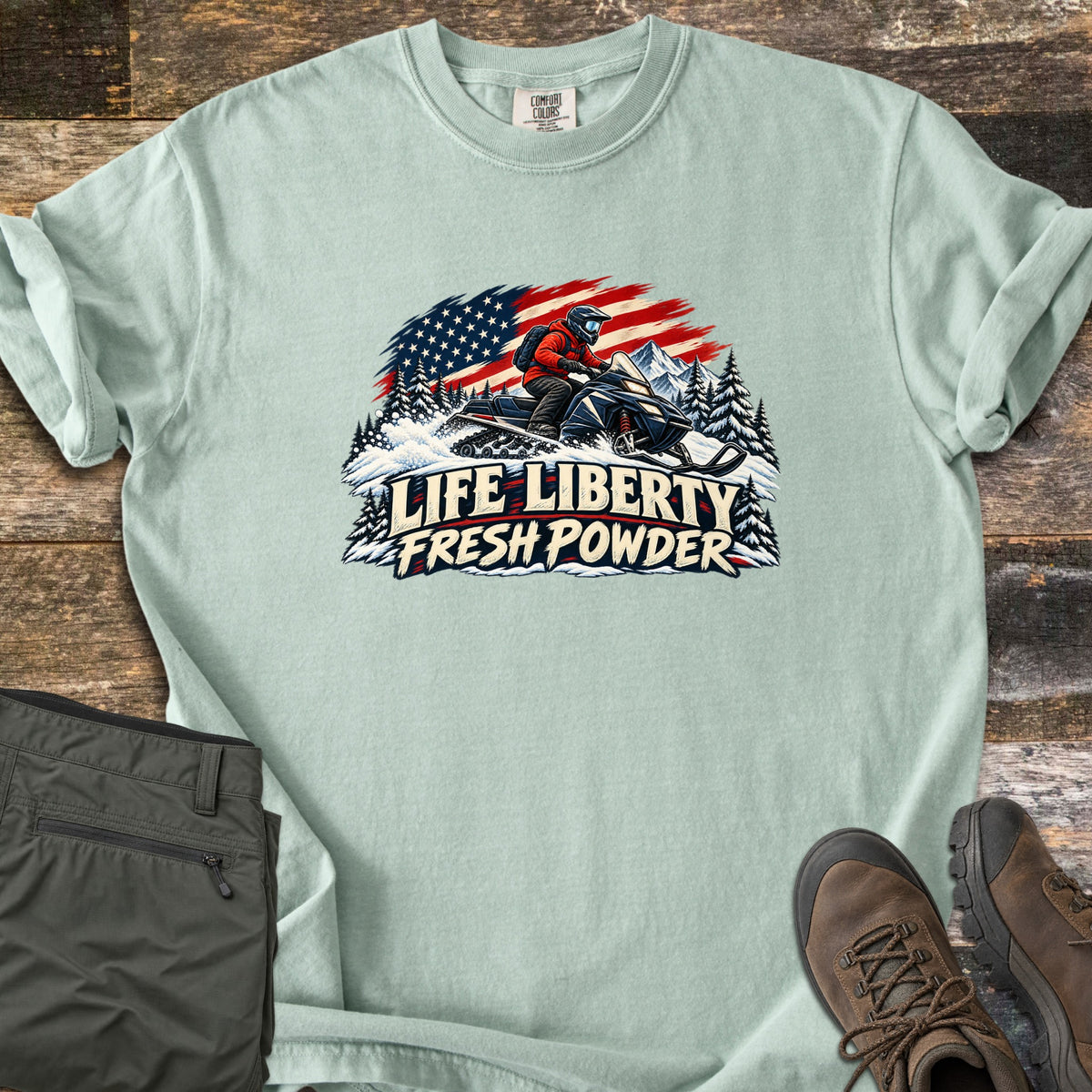 Life Liberty Fresh Powder Comfort Colors T-shirt in Bay, laid flat. A rider in a red jacket and helmet drives a snowmobile through snowy pines past mountains and a waving American flag, above the words Life Liberty Fresh Powder. Gray pants and brown hiking boots lie on weathered wood.