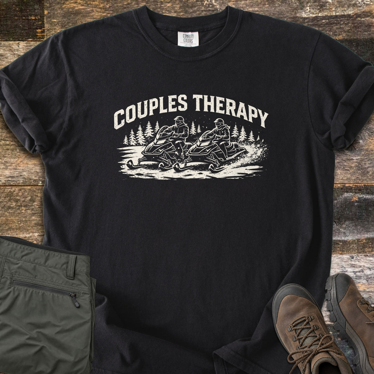 Couples Therapy Snowmobile Comfort Colors T-shirt in Black, laid flat. The words COUPLES THERAPY arch in white over two riders on snowmobiles kicking up snow among pine trees. Olive cargo pants and brown hiking boots lie beside it on dark weathered wood.