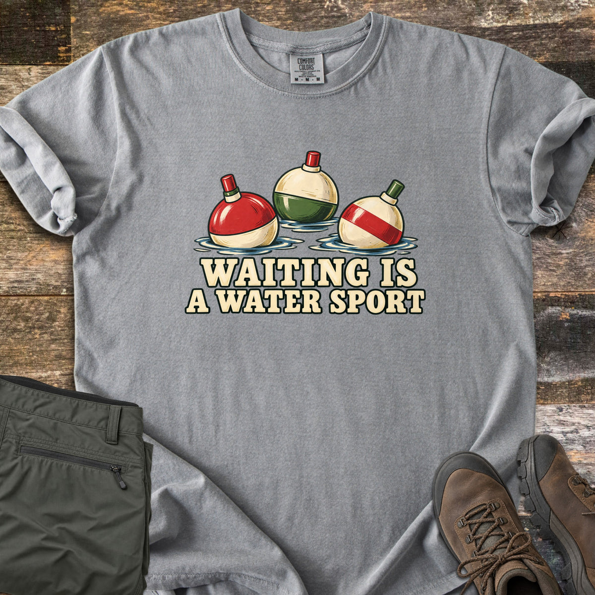 Waiting Is a Water Sport Comfort Colors T-shirt in Grey, laid flat. Three red, green and cream fishing bobbers float in water above the words Waiting Is a Water Sport. The shirt lies on weathered wood beside folded olive pants and brown hiking boots.
