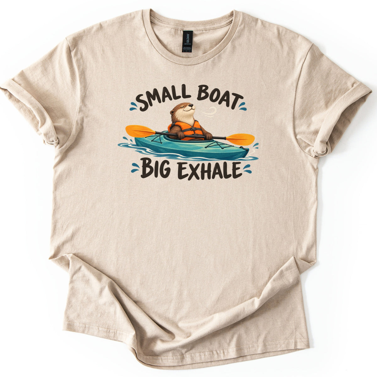 Small Boat Big Exhale Comfort Colors T-shirt in Sand, laid flat. A smiling otter in an orange life vest paddles a teal kayak with an orange paddle, between the words Small Boat and Big Exhale. The shirt has rolled sleeves on a white background.