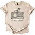 Chickadee FM Comfort Colors T-shirt in Sand, laid flat. A sketched chickadee sings from atop a vintage radio above the words CHICKADEE FM and SAME TWO NOTES ALL DAY, printed in black. The shirt has rolled sleeves and lies on a white background.