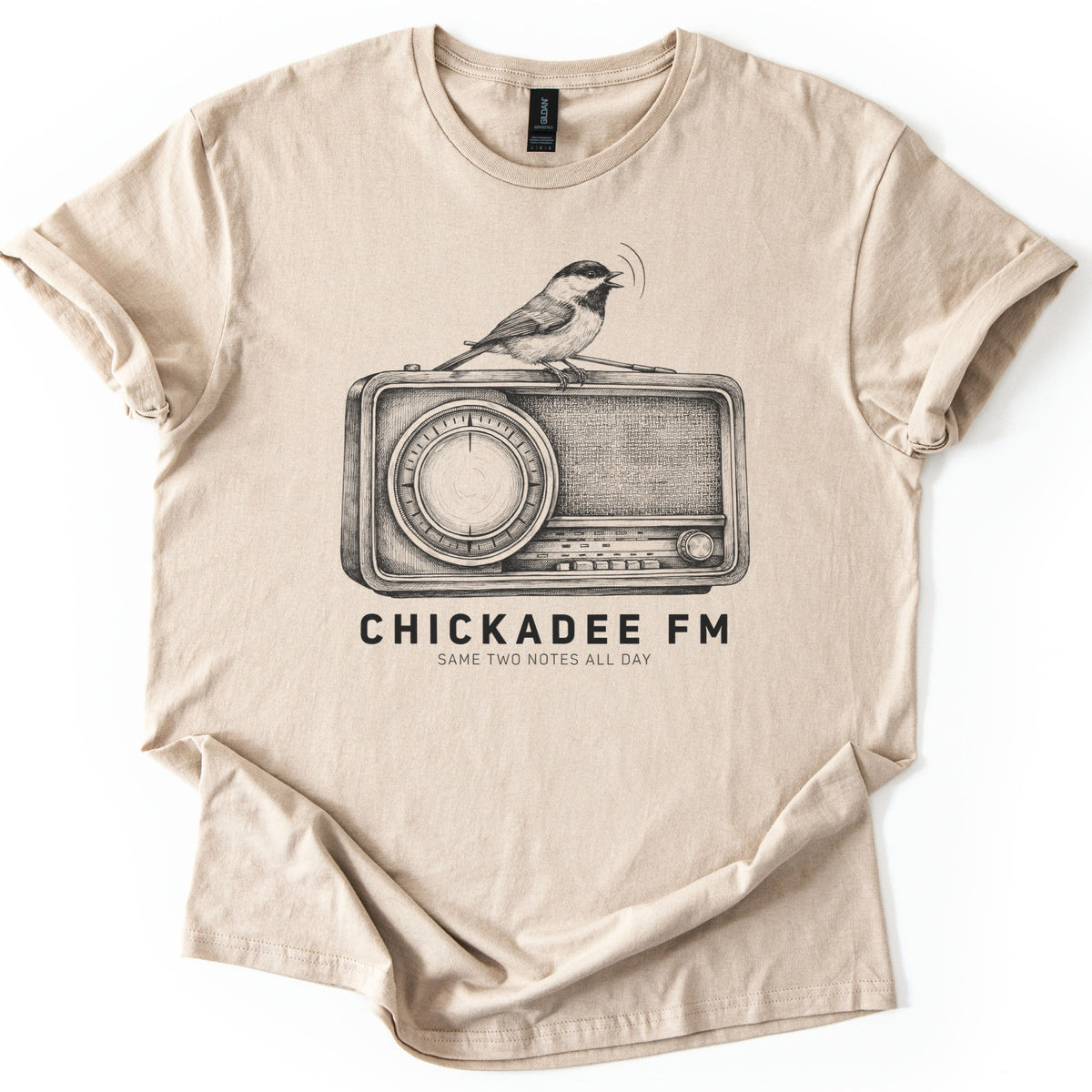 Chickadee FM Comfort Colors T-shirt in Sand, laid flat. A sketched chickadee sings from atop a vintage radio above the words CHICKADEE FM and SAME TWO NOTES ALL DAY, printed in black. The shirt has rolled sleeves and lies on a white background.