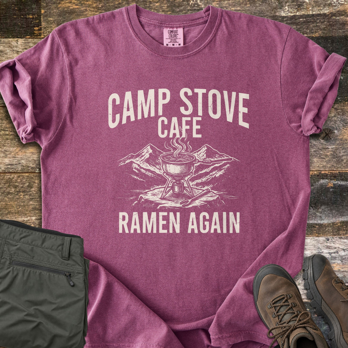 Camp Stove Cafe Comfort Colors T-shirt in Berry, laid flat. Printed in cream: a steaming pot on a camp stove atop a rock with mountains behind, under the words Camp Stove Cafe and above Ramen Again. Sleeves rolled, on weathered wood with green pants and hiking boots.