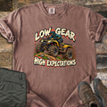 Low Gear High Expectations Comfort Colors T-shirt in Espresso, laid flat. A rider in a teal helmet drives a muddy yellow ATV over a dirt mound, between the words Low Gear and High Expectations. Folded green cargo pants and brown hiking boots lie on weathered wood.
