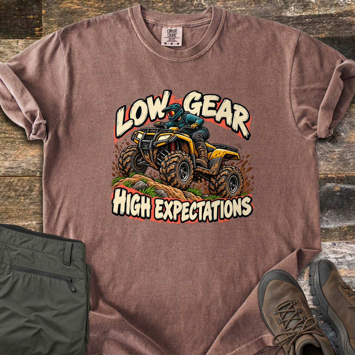 Low Gear High Expectations Comfort Colors T-shirt in Espresso, laid flat. A rider in a teal helmet drives a muddy yellow ATV over a dirt mound, between the words Low Gear and High Expectations. Folded green cargo pants and brown hiking boots lie on weathered wood.