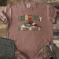 Snow Day Fully Booked Comfort Colors T-shirt in Espresso, laid flat. A rider in an orange jacket and helmet speeds on an orange and white snowmobile through snow between the words Snow Day and Fully Booked in green, cream and plaid letters. Olive pants and brown boots sit on a wood floor.