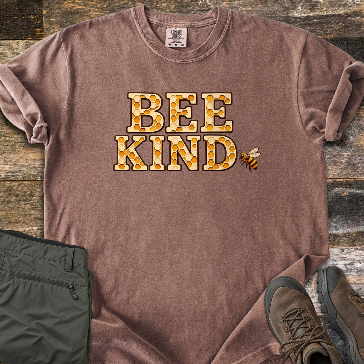 Bee Kind Comfort Colors T-shirt in Espresso, laid flat. The words BEE KIND in large letters filled with a golden honeycomb pattern, with a small bee beside them. Sleeves are rolled, on a weathered wood floor beside folded olive cargo pants and brown hiking boots.