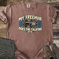 Freehub Does the Talking Comfort Colors T-shirt in Espresso, laid flat. A black and cream bike rear hub with blue sound waves sits between the words My Freehub and Does the Talking, with MTB below. Olive hiking pants and brown boots lie beside it on weathered wood.