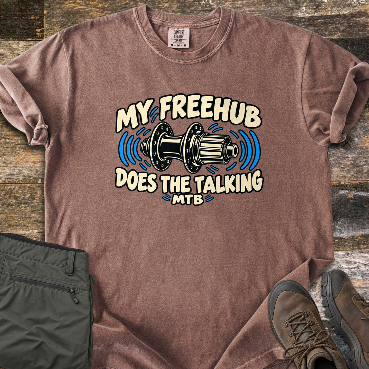 Freehub Does the Talking Comfort Colors T-shirt in Espresso, laid flat. A black and cream bike rear hub with blue sound waves sits between the words My Freehub and Does the Talking, with MTB below. Olive hiking pants and brown boots lie beside it on weathered wood.
