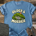 Older & Mossier Comfort Colors T-shirt in Blue Jean, laid flat. A smiling gray rock covered in green moss and wearing a red polka-dot party hat sits between the words Older & Mossier in cream letters. Folded olive hiking pants and brown hiking boots lie on weathered wood.
