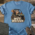 Nut Driver Comfort Colors T-shirt in Blue Jean, laid flat. A cartoon squirrel grins while driving a muddy camo side-by-side with an acorn on the hood, above the words Nut Driver. Olive cargo pants and brown hiking boots sit beside it on weathered wood.
