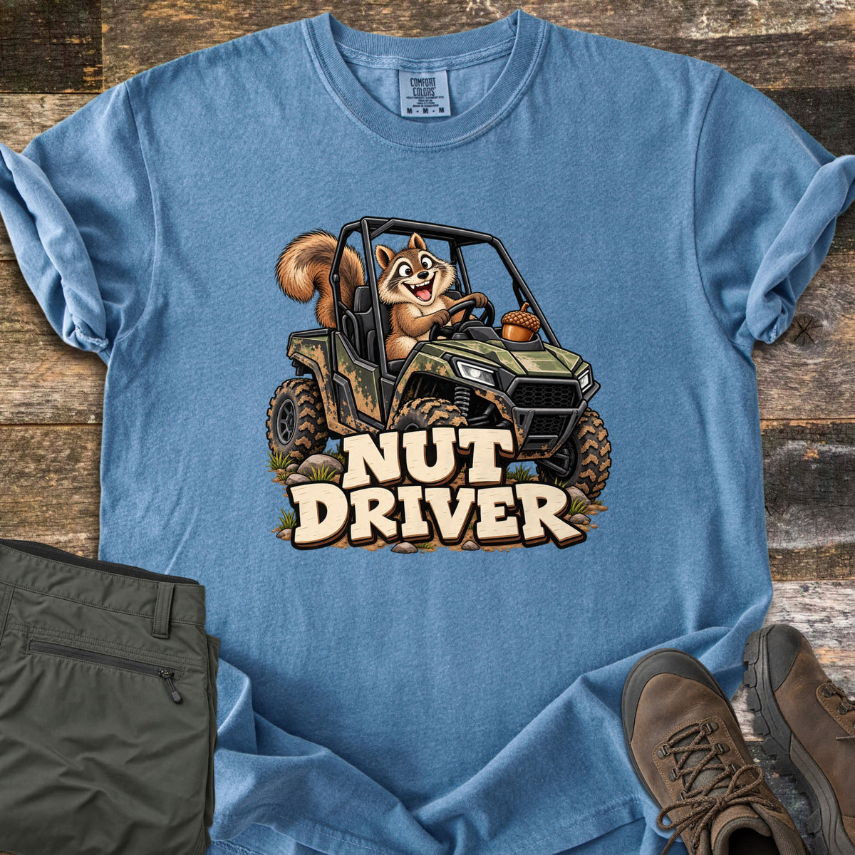 Nut Driver Comfort Colors T-shirt in Blue Jean, laid flat. A cartoon squirrel grins while driving a muddy camo side-by-side with an acorn on the hood, above the words Nut Driver. Olive cargo pants and brown hiking boots sit beside it on weathered wood.