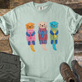 Patchwork Otter Comfort Colors T-shirt in Bay, laid flat. Three patchwork otters in orange, peach and turquoise float on their backs, each in a gingham or striped body hugging a small fish, with X-stitched eyes. Olive pants and brown hiking boots sit on a wood floor.
