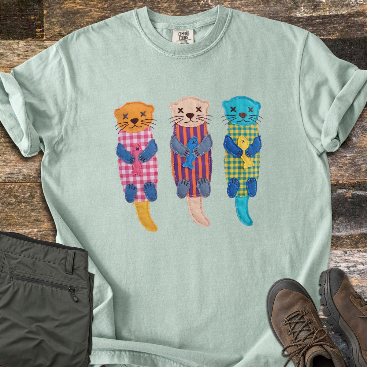 Patchwork Otter Comfort Colors T-shirt in Bay, laid flat. Three patchwork otters in orange, peach and turquoise float on their backs, each in a gingham or striped body hugging a small fish, with X-stitched eyes. Olive pants and brown hiking boots sit on a wood floor.