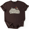 Fresh Corduroy Comfort Colors T-shirt in Dark Chocolate, laid flat. A cream and red illustration of a rider on a snowmobile cresting a groomed slope with curving corduroy lines and two pine trees, above the words Fresh Corduroy. Sleeves are rolled on a white background.