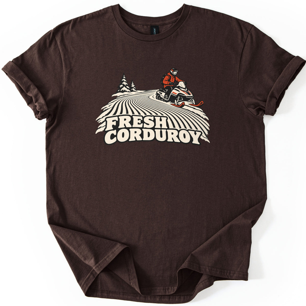 Fresh Corduroy Comfort Colors T-shirt in Dark Chocolate, laid flat. A cream and red illustration of a rider on a snowmobile cresting a groomed slope with curving corduroy lines and two pine trees, above the words Fresh Corduroy. Sleeves are rolled on a white background.