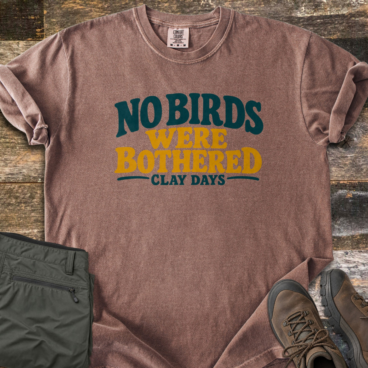 No Birds Were Bothered Comfort Colors T-shirt in Espresso, laid flat. Printed on the chest in dark teal and mustard yellow lettering: No Birds Were Bothered, with Clay Days below. Sleeves rolled, on weathered wood with olive cargo pants and brown hiking boots.