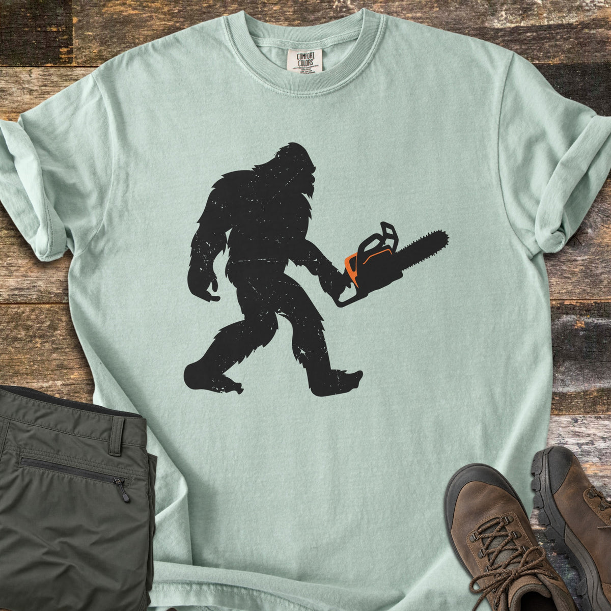 Lumber Squatch Comfort Colors T-shirt in Bay, laid flat. A distressed black silhouette of Bigfoot walks while carrying a chainsaw with orange accents. The sleeves are rolled, with folded olive pants and brown hiking boots on rustic wood planks.