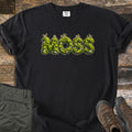 Moss Comfort Colors T-shirt in Black, laid flat. The word MOSS is printed in letters made of green moss with small stones and sprouting stalks. The shirt lies on weathered wood planks beside folded olive hiking pants and brown hiking boots.