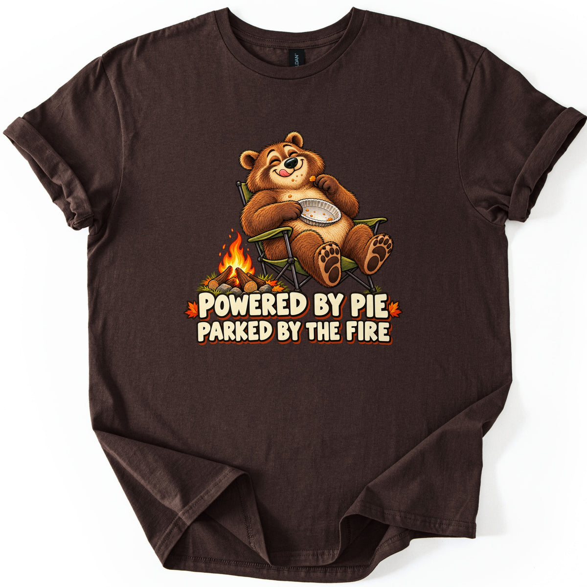 Powered by Pie Parked by the Fire Comfort Colors T-shirt in Dark Chocolate, laid flat. A smiling cartoon bear licks its lips in a green camp chair, holding an empty pie tin beside a campfire, above the words Powered by Pie Parked by the Fire. The shirt has rolled sleeves on a white background.