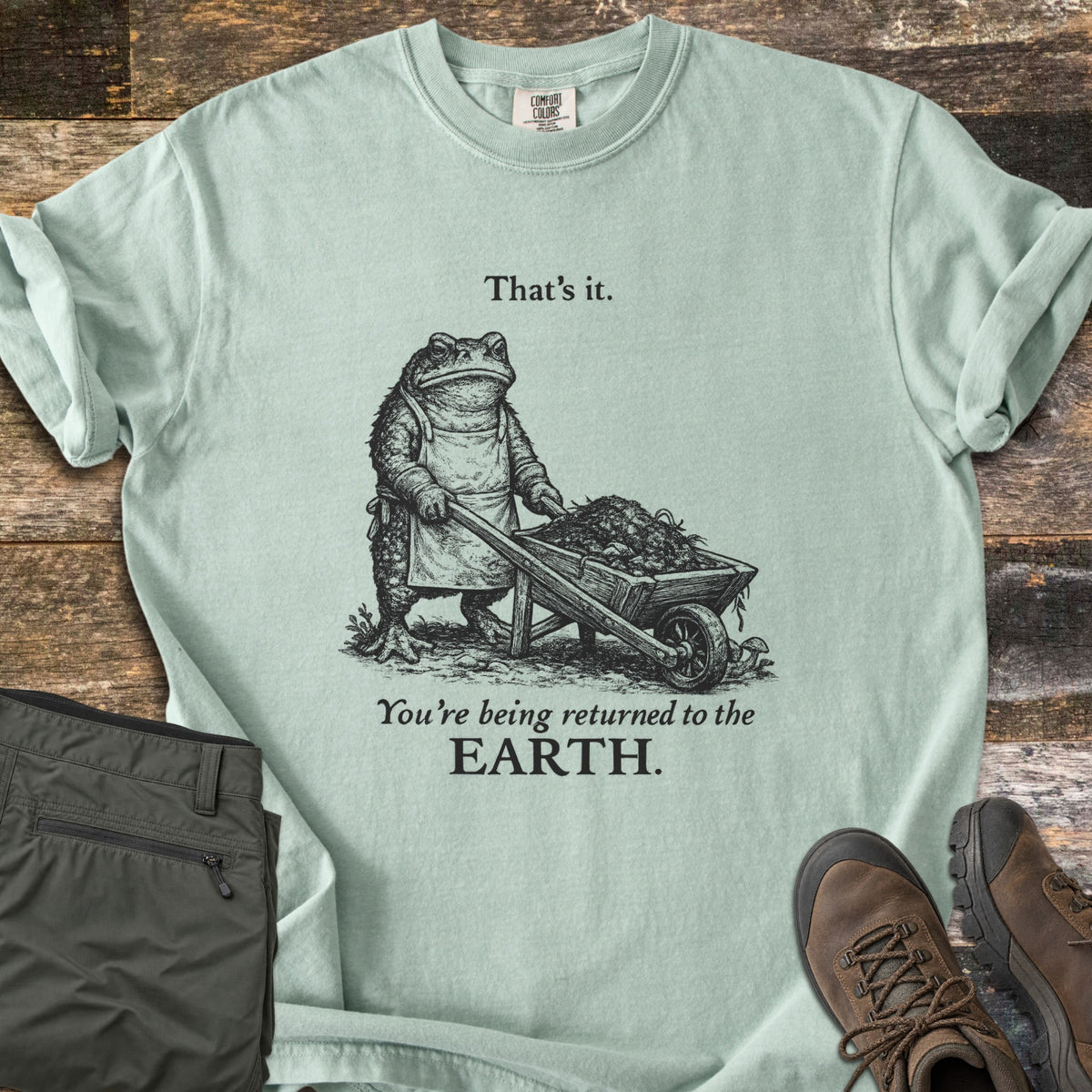 Toad Compost Comfort Colors T-shirt in Bay, laid flat. A black-ink toad in an apron pushes a wheelbarrow of compost between the words That's it. and You're being returned to the EARTH. Folded olive pants and brown hiking boots lie beside it on weathered wood.