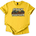 Stuck Together Comfort Colors T-shirt in Daisy, laid flat. Two mud-covered green ATVs joined by an orange tow strap sit between the words Stuck Together in distressed cream letters. The shirt has cuffed sleeves and lies on a white background.