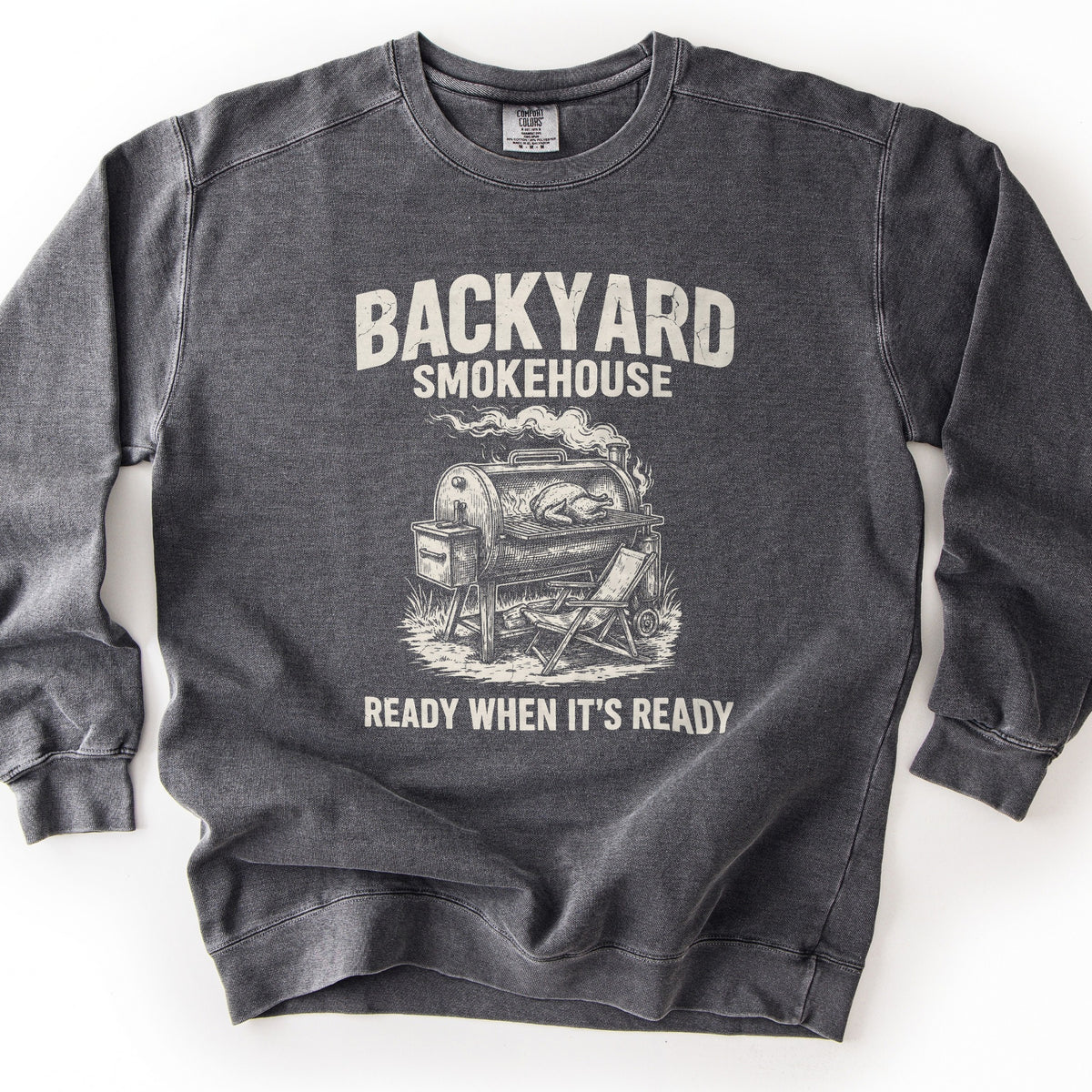 Backyard Smokehouse Comfort Colors crewneck sweatshirt in Pepper, laid flat. Large words Backyard Smokehouse sit above a sketch of a barrel smoker with a turkey on the grate, smoke rising, and a folding chair beside it, over the words Ready When It's Ready. Shown on a white background.