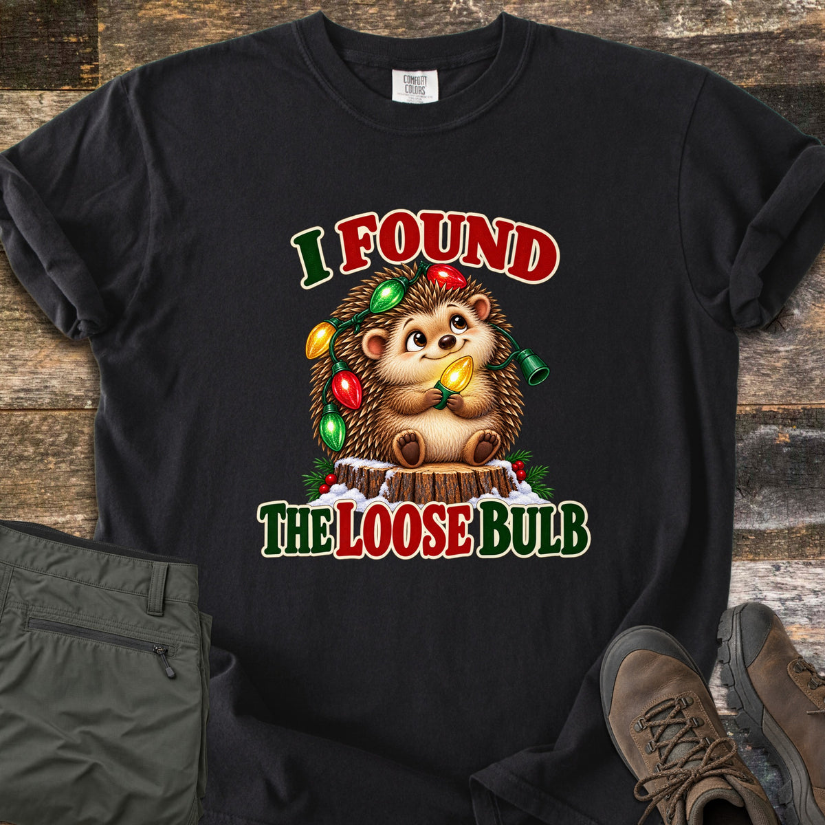 I Found The Loose Bulb Comfort Colors T-shirt in Black, laid flat. A cartoon hedgehog sits on a snowy tree stump, wrapped in red, green and yellow Christmas lights and holding a glowing bulb, between the words I Found and The Loose Bulb. Olive pants and brown boots lie on a wood floor.