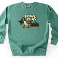 Fowl Play Comfort Colors crewneck sweatshirt in Light Green, laid flat. A cartoon Canada goose carries a green motocross helmet in its beak past a green four-wheeler and pine trees, under the words FOWL PLAY in cream letters. The sweatshirt lies on a white background.