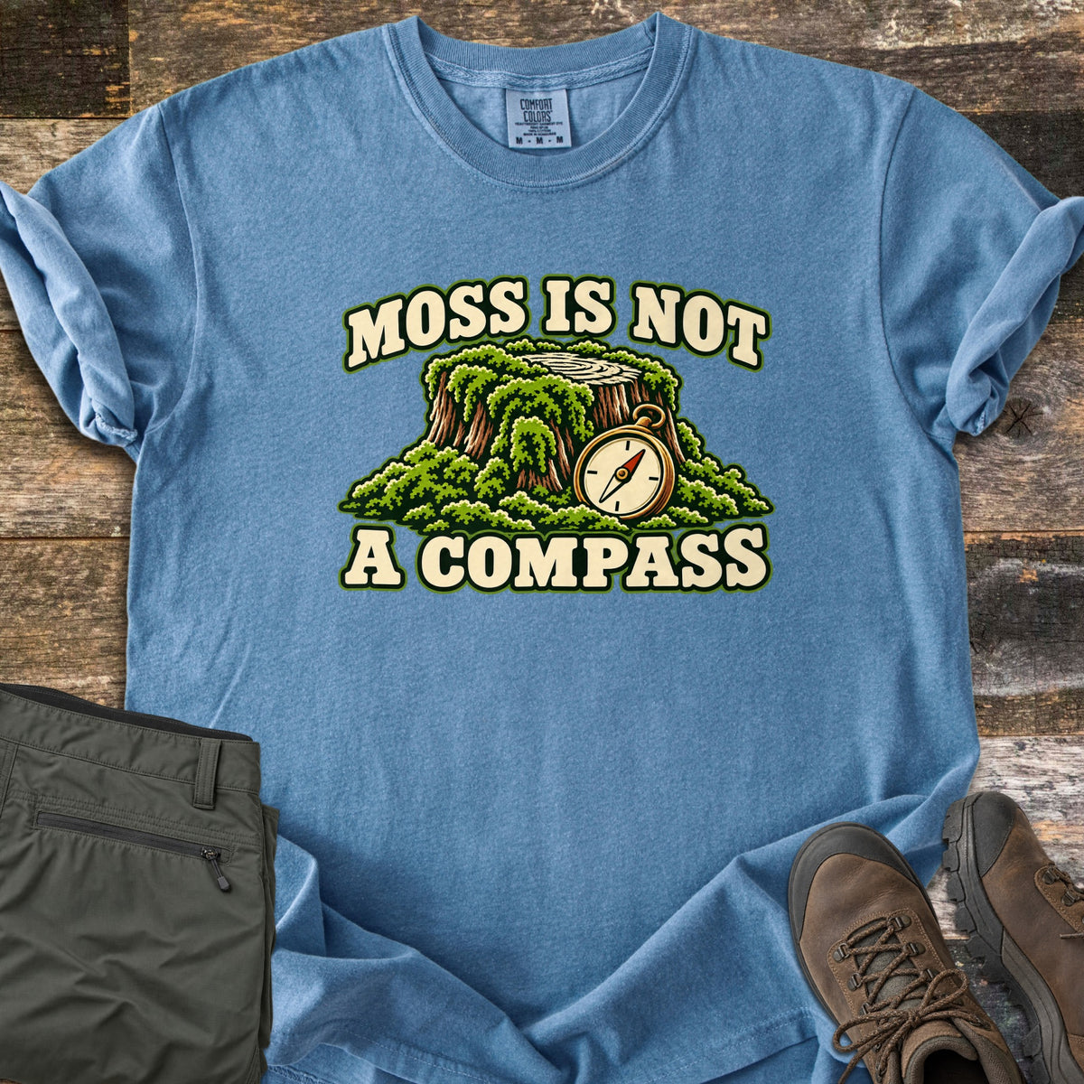 Moss Is Not a Compass Comfort Colors T-shirt in Blue Jean, laid flat. A moss-covered tree stump with a brass compass leaning on it, between the words MOSS IS NOT and A COMPASS. Folded olive hiking pants and brown hiking boots sit beside it on weathered wood planks.