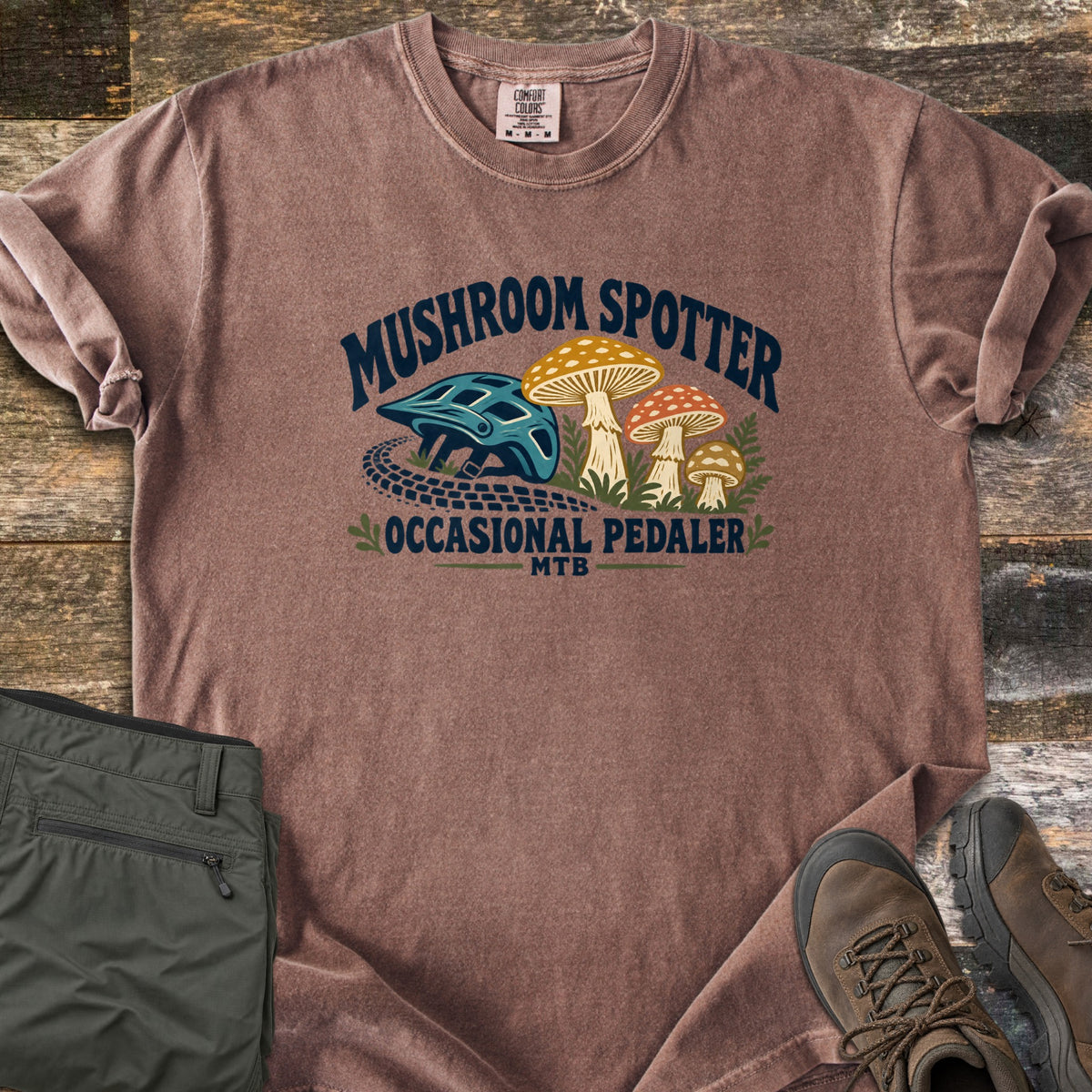 Mushroom Spotter Occasional Pedaler Comfort Colors T-shirt in Espresso, laid flat. A teal bike helmet, a tire track and three spotted mushrooms sit under the arched words Mushroom Spotter, with Occasional Pedaler MTB below. It lies on wood planks with olive pants and brown hiking boots.