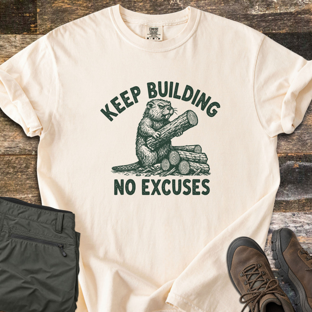 Keep Building Beaver Comfort Colors T-shirt in Ivory, laid flat. A sketch-style beaver hugs a log beside a stack of logs, printed in dark green with the words Keep Building above and No Excuses below. Olive cargo pants and brown hiking boots lie beside it on weathered wood.